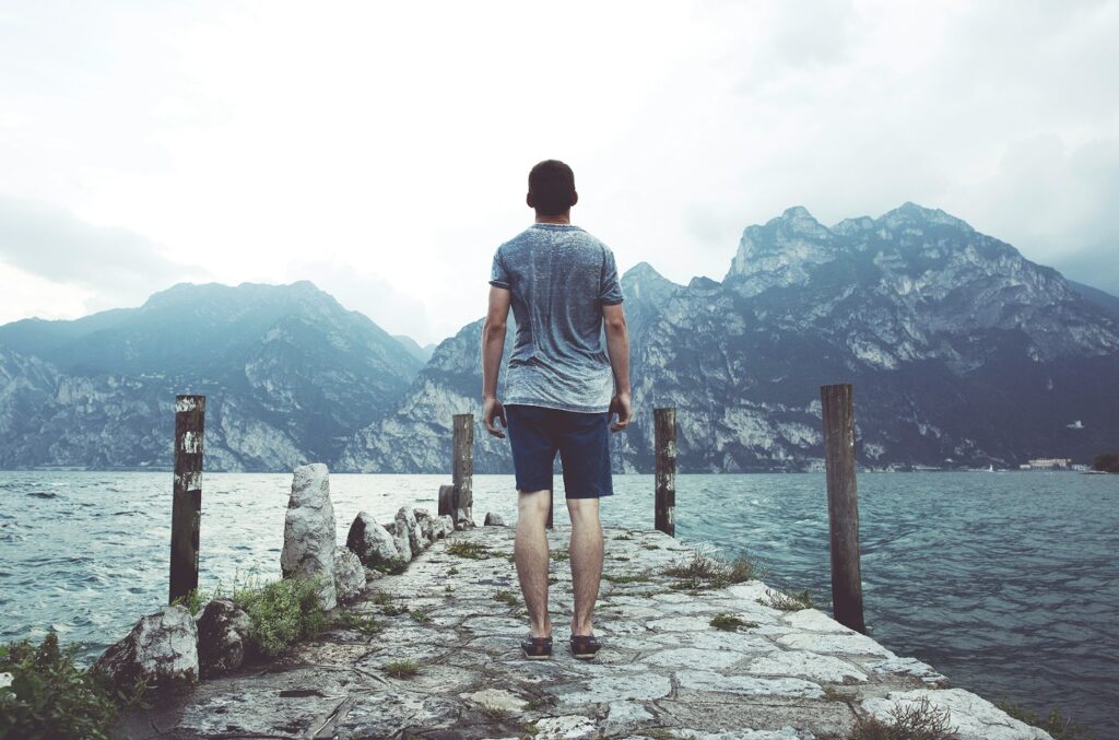 man standing on gray concrete dock facing body of water and mountains at daytime